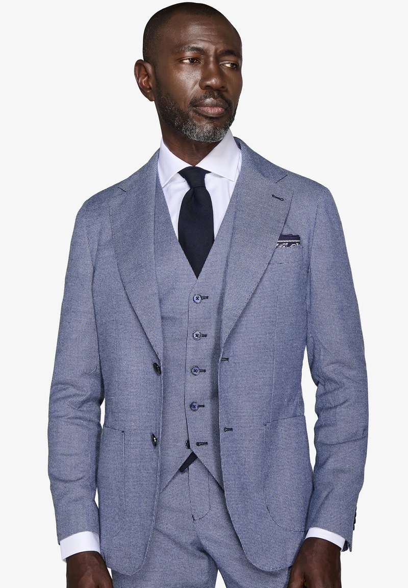 Completo a tre pezzi blu a quadri realizzato in tessuto testurizzato. Presenta rever a punta, gilet con bottone e cravatta scura. Il fazzoletto da taschino aggiunge un tocco di dettaglio.