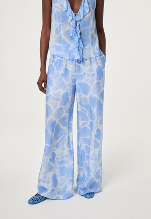 Vrouw draagt blauw-witte tie-dye wijde broek met zakken, bijpassende gerimpelde mouwloze top en blauwe geperforeerde instapschoenen.