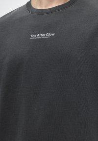 Σκούρο γκρι φανελάκι με λευκό κείμενο στο κέντρο του στήθους που γράφει «The After Glow sending energy into space.»