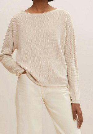 Femme portant un pull en maille texturée beige clair à manches longues et un pantalon de couleur crème, la main reposant dans la poche, sur un fond uni.