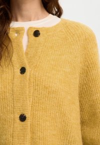 Femme portant un cardigan en maille jaune doux avec des boutons foncés sur un haut blanc à col rond, visible des épaules jusqu'au milieu de la poitrine.