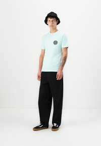 Lyseblå kortermet t-skjorte med svart rund grafikk, kombinert med svarte vide jeans og svarte sneakers med hvite detaljer. Svart bøttehatt.