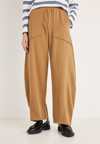 Bruine wijde broek gemaakt van zachte stof, met grote voorzakken en een losse pasvorm. Gecombineerd met zwarte schoenen en een gestreept shirt met lange mouwen.