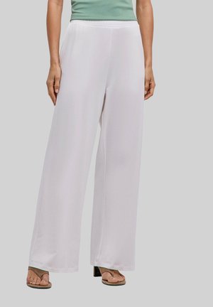 Persona che indossa pantaloni bianchi larghi a gamba larga, top verde senza maniche e sandali con tacco marroni, in piedi contro uno sfondo semplice.