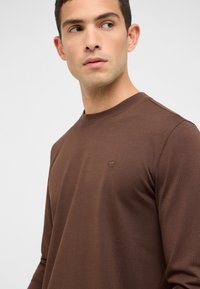 Braunes Langarm-T-Shirt aus weichem Stoff, mit rundem Ausschnitt und einem kleinen Logo. Glatte Textur mit figurbetontem Schnitt.