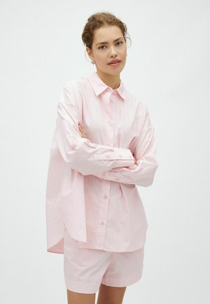 Young woman with curly hair wears oversized pale pink button-up shirt and matching shorts, standing with arms crossed against plain background.