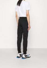 Schwarze Jogginghose mit einem strukturierten Bund, ausgestattet mit einer Gesäßtasche mit grünen Akzenten. Kombiniert mit einem weißen T-Shirt mit kurzen Ärmeln und sportlichen Schuhen.