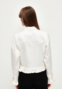Giacca bianca in satin con un design cropped, orlo e polsini con volant e una leggera lucentezza. Presenta un collo classico e dettagli di cucitura pulita.