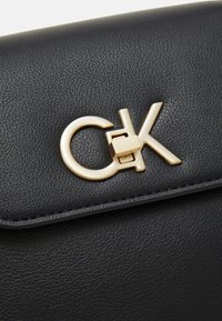 Rabattant de sac en cuir noir texturé avec une boucle dorée au logo Calvin Klein centrée sur l’avant.