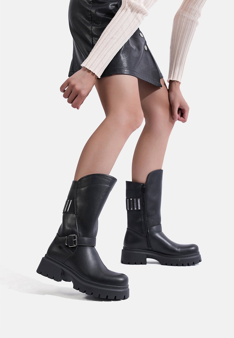 Persona che indossa stivali neri in pelle fino a metà polpaccio con suole spesse e una minigonna nera in pelle, in piedi contro uno sfondo bianco semplice.