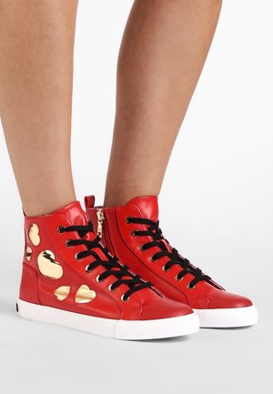 Czerwone sneakersy za kostkę z czarnymi sznurowadłami, złote naszywki w kształcie serca po bokach, białe podeszwy, noszone na gołych nogach na białym tle.