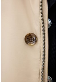 Beige outerwear with a textured finish, featuring a large transparent button with floral design and a silver zipper along the side.