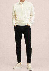 Suéter de media cremallera en beige claro con ribete acanalado, combinado con jeans negros ajustados con desgastes en las rodillas y zapatillas ligeras.