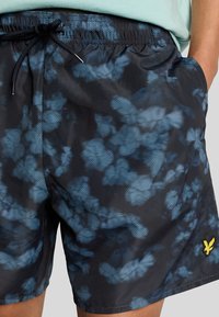 Pantaloni da bagno neri con un motivo astratto blu, vita con cordino e tasche laterali. Accento distintivo del logo giallo sull'orlo.