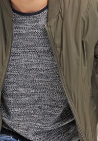 Bomber bunda olivově zelené barvy s žebrovaným límcem a zipem, nošená přes texturovaný šedý svetr. Ve výšce pasu jsou vidět casual džíny z denimu.