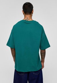Teal oversized t-shirt van katoen, met een ronde hals en korte mouwen. De achterkant toont een zichtbare tatoeage en heeft een relaxte pasvorm.