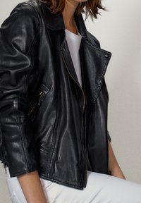 Personne portant une veste en cuir noire sur une chemise blanche et un pantalon blanc sur un fond clair uni, cadrée de la moitié du visage jusqu’à la moitié de la cuisse.