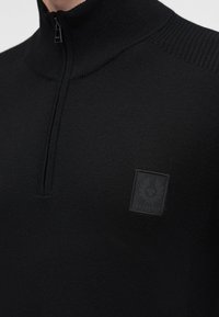 Pull en maille noire avec un col à fermeture éclair jusqu’au quart et un petit logo carré sur la poitrine.