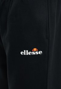 Μαύρο ύφασμα με μικρό λευκό λογότυπο «ellesse» και πορτοκαλοκόκκινο ημικύκλιο σχεδιασμένο πάνω από το κείμενο, στο κέντρο του ρούχου.