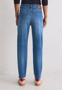 Personne portant un jean bleu et des baskets blanches avec des accents rouges, se tenant contre un mur léger uni, vu de derrière.