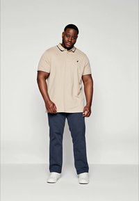Polo beige avec un col et deux boutons, arborant un petit logo sur la poitrine ; associé à un pantalon bleu marine et des baskets blanches.