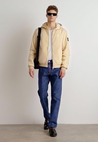 Svetlo rumena puffer jakna s kapuco, bela majica, modri jeans in črne čevlje. Model nosi črne sončne očala in nosi črno torbo.
