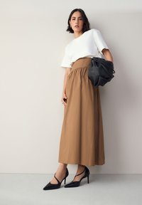 Brązowa spódnica sięgająca kostek z marszczoną talią, zestawiona z białym, krótkim topem i czarnymi butami na obcasie. Dodatki obejmują czarną torebkę.