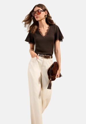 Frau trägt braunes Spitzen-V-Neck-Oberteil, cremefarbene Hosen mit weitem Bein, übergroße Sonnenbrille, goldene Ohrringe, hält braune Clutch mit Quasten.
