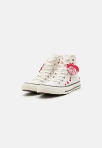 White high-top sneakers made of canvas, featuring red heart accents and a red rose tag. Black stripes along the sole for contrast.