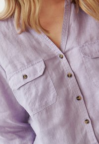 Lichtpaarse linnen shirt met een V-hals, twee voorkruiken en vijf donkere knopen. Het heeft een ontspannen pasvorm en zachte, gestructureerde stof.