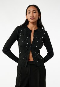 Cardigan nero a coste con maniche lunghe, scollatura tonda, chiusura frontale con bottoni e dettagli in perle su tutta la superficie. Il materiale sembra morbido e testurizzato.