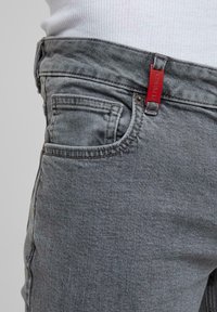 Gros plan sur une poche de jean en denim gris avec une étiquette rouge sur la boucle de ceinture et une partie d'un t-shirt blanc côtelé rentré.