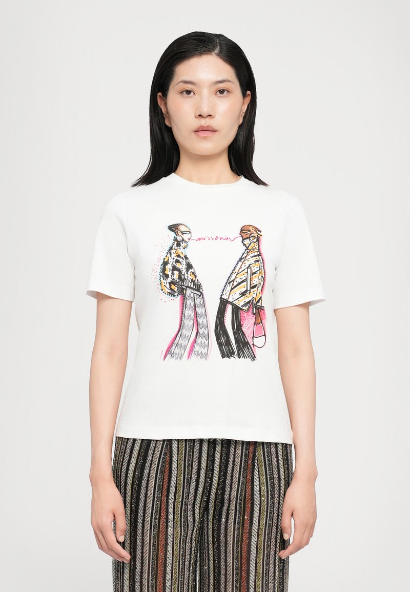 T-shirt en coton blanc avec un design graphique coloré représentant deux personnages, avec des lignes et des motifs audacieux. Le modèle porte un pantalon rayé.