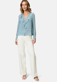 Blouse bleu clair avec un devant froncé et des bordures blanches, associée à un pantalon large crème. Stylisée avec des talons ouverts blancs.