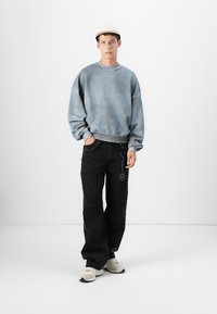 Moleton oversized azul desbotado, jeans pretos de perna larga com detalhes desgastados e embellishments, combinados com ténis beges e um boné.