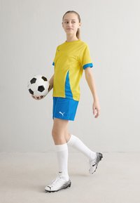 Ungdomsfotbollsdräkt med gul tröja med blå detaljer, blå shorts, vita strumpor och en svartvit fotboll.