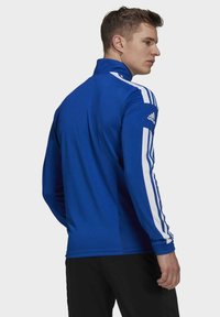 Blauwe sportjas met een hoge kraag, witte strepen langs de mouwen en een Adidas-logo op de linkermouw. Glad textuur van de stof.