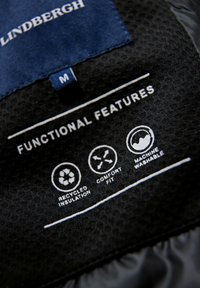 Svart tygmärke med texten "FUNKTIONELLA FUNKTIONER", ikoner för återvunnen isolering, bekväm passform och maskintvättbar; inkluderar varumärket "Lindbergh" och storlek "M."