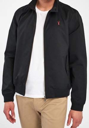 Homme portant une veste zippée noire avec un logo de cerf rouge sur la poitrine, un t-shirt blanc et un pantalon beige, montré du cou aux hanches.
