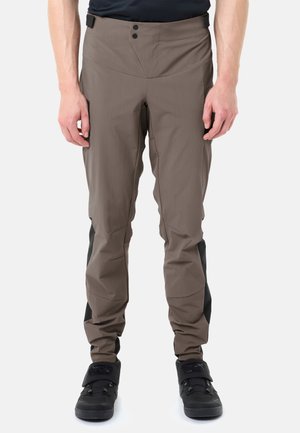 Mann trägt braune Outdoor-Hose mit verstärkten schwarzen Einsätzen an den Waden und schwarzen High-Top-Schuhen, steht vor weißem Hintergrund.