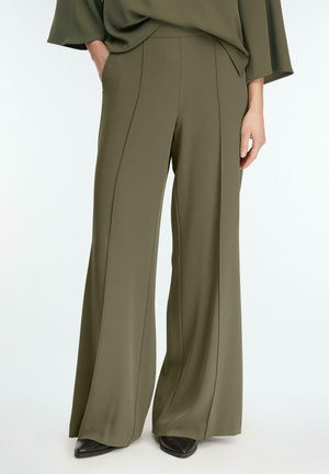 Osoba ubrana w oliwkowozielone szerokie spodnie i dopasowaną górę, z jedną ręką w kieszeni i widocznymi czarnymi, spiczastymi butami.