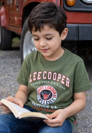 Garçon en t-shirt vert Lee Cooper lisant un livre assis sur du gravier à côté d'un véhicule rouge.