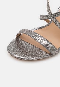 Silverfärgade metallsandaler med en strukturerad yta, öppen tå och korskorsande remmar. Sulan är ljusbrun med en slät yta.