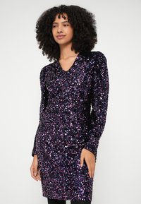 Γυναίκα με κυματιστά μαλλιά που φοράει μαύρο μίνι φόρεμα με μακριά μανίκια καλυμμένο με μωβ και μπλε παγιέτες, στέκεται μπροστά σε απλό φωτεινό φόντο.