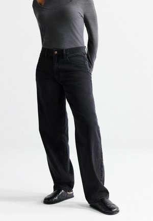 Person iført sorte bredbenede jeans, en tætsiddende grå langærmet top og sorte vævede slip-on sko, står med hænderne bag ryggen.