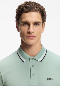 Lysegrøn poloshirt med tekstureret stof, tre knapper og en kontrastfarvet sort-og-hvid stribet krave; har en lille 'BOSS'-logo.