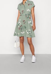 Blumenkleid mit kurzen Ärmeln in Grün, mit Knopfleiste, tailliertem Bund und fließendem Rock. Kombiniert mit weißen Sneakers und einer kleinen Handtasche.