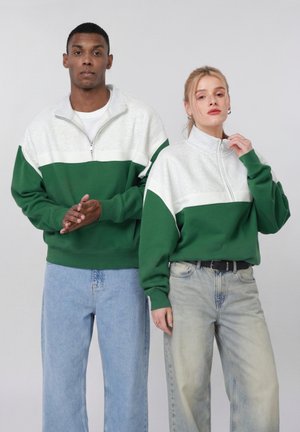 Mężczyzna i kobieta stoją obok siebie, ubrani w zielono-białe bluzy z suwakiem do połowy, oraz jasnoniebieskie dżinsy, na jednolitym tle.