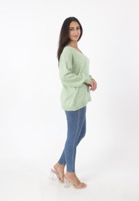 Lichtgroene gebreide trui met een V-hals en lange mouwen, gecombineerd met blauwe skinny jeans en doorzichtige haksandalen.