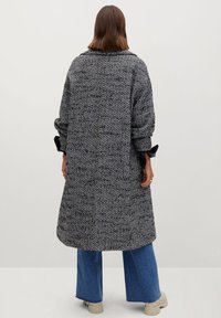 Person, der einen langen schwarz-weißen Fischgrätenmantel, blaue weite Jeans und beige Schuhe trägt, steht vor einem schlichten hellen Hintergrund.